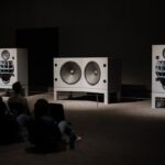 Drei große Lautsprecher in einer abgedunkelten Ausstellung mit sitzendem Publikum